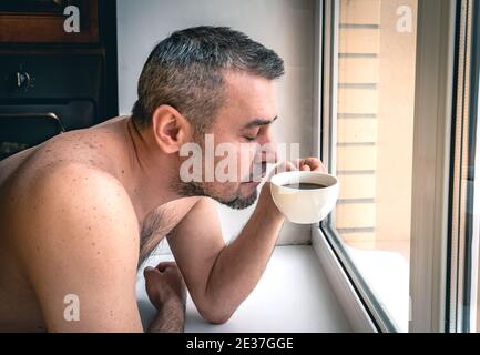 Nahaufnahme eines bärtigen Mannes, der Kaffee trinkt. Porträt eines Mannes, der vor einem sonnendurchfluteten Fenster eine Tasse Tee genießt. Stockfoto