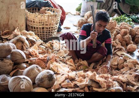 Ein junger Mann im traditionellen Longyi sitzt und hackt Kokosnüsse auf einem lokalen Obstmarkt in der Nähe von Yangon, Myanmar Stockfoto