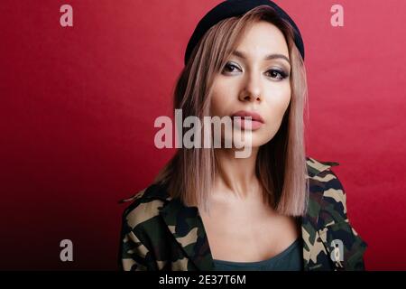 Nahaufnahme Porträt der blonden Frau vor rotem Hintergrund. Frau in schwarzer Mütze und Militärjacke Stockfoto