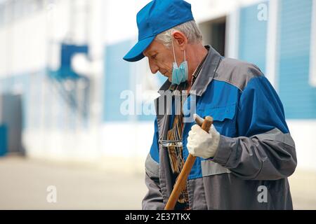 Dies ist ein älterer Hausmeister mit einem Besen in einer medizinischen Maske auf der Straße fegen das Gebiet. Stockfoto