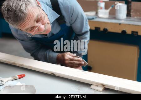 Zimmermannsarbeit in der Schreinerei. Ein älterer männlicher Tischler untersucht sorgfältig ein Holzbrett. Stockfoto