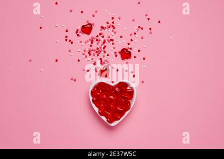 Weiße Keramik herzförmige Schüssel mit Spritzer von roten herzförmigen Konfetti und kleine dekorative Herzen auf rosa Hintergrund. Valentinstag-Konzept. Oben Stockfoto