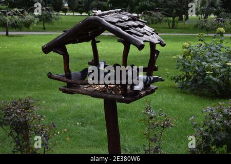 Handgefertigter Vogelfutterhäuschen mit Vögeln. Vogelfutterhäuschen aus Holz in Form eines Hauses mit einem Dach, an einem Holzpfosten in einem Park zwischen Bäumen in der Natur befestigt Stockfoto
