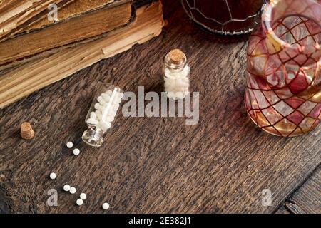 Eine Flasche homöopathischer Pillen verschüttet auf einem Holztisch, mit alten Büchern im Hintergrund Stockfoto