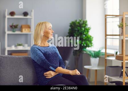 Nachdenklich lächelnde ältere Frau, die aus dem Fenster blickt und auf dem Sofa im gemütlichen Wohnzimmer sitzt. Stockfoto