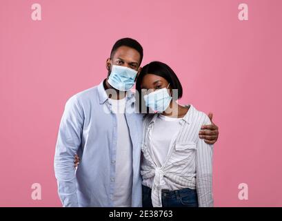 Valentinstag während des Coronavirus-Ausbruchs. Unglücklich schwarzes Paar in Gesichtsmasken Umarmung auf rosa Hintergrund Stockfoto