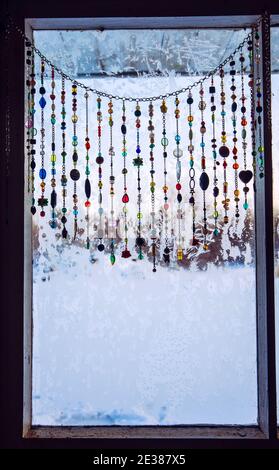 Handgefertigter Sonnenfänger mit Kristallen und Perlenfenstervorhängen, die an winterfrostigen Fenstern hängen. Stockfoto