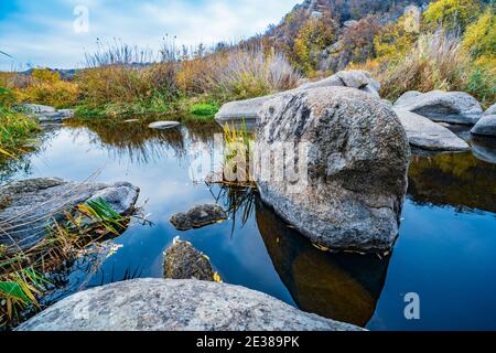 Ein schneller, flacher, sauberer Bach fließt zwischen glatten nassen großen Steinen, umgeben von hohen trockenen Klumpen, die im Wind im malerischen Ukraine schwanken Stockfoto