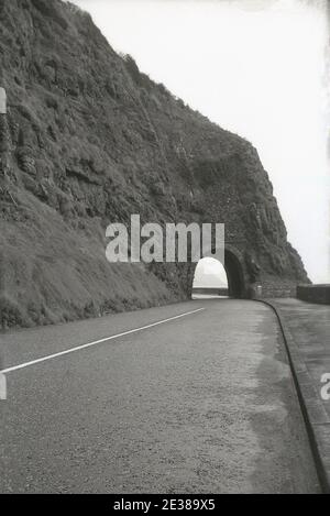 1950er Jahre, historisch, in Larne, am Anfang der Causeway Küstenstraße (A2), geht die Straße unter dem Blackcave Tunnel oder "Black Arch", eine enge Öffnung oder Tunnel aus der überhängenden Klippe geschnitten, Larne, Antrim, Nordirland. Es war die Idee des Bauingenieurs William bald, der den Spielraum für den Bau einer Straße entlang der Kante der Klippe sah, die zwischen 1832 und 1842 stattfand und eine große Leistung für seine Zeit war. Die Straße blieb bis in die späten 1960er Jahre weitgehend unverändert. Stockfoto