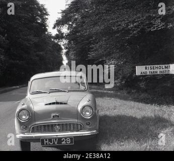 1950er Jahre, historisch, ein Austin Auto geparkt auf dem Gras neben einer Straße, gegenüber einem Schild zu Riseholme & Farm Institute, eine landwirtschaftliche Hochschule im Jahr 1949 gegründet. Die Halle wurde von Lincolnshire Education Committee von der Wilson-Familie, erworben hatte es im Jahr 1870. 1966 wurde Riseholme das Lindsey College of Agriculture. 1980, fusioniert mit Kesteven Agricultural College und Holbeach Agricultural Center, um das Lincolnshire College of Agriculture and Horticulture zu bilden. Im Jahr 1994 wurde es Teil der De Montfort University, Leicester. Später wurde es an die University of Lincoln übertragen. Stockfoto