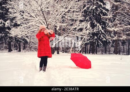 Eine Frau mit rotem Regenschirm macht Fotos und Videos auf ihrem Handy in einem Winterwald. Lächelnde Frau in roter Jacke mit dem Telefon im Park Stockfoto