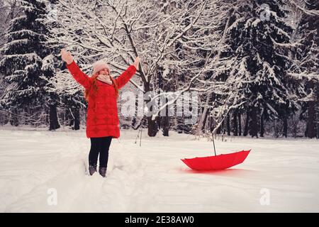 Lächelnde Frau mit einem roten Schirm genießt weißen Schnee in einem Winterwald. Eine glückliche Frau in roten Kleidern steht im Park. Stockfoto