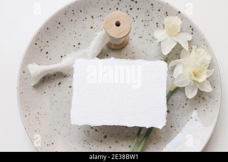 Frühling Schreibwaren Stillleben. Leere Visitenkarte, Einladung. Osterszene mit Narzissen, Narzissen Blumen auf Porzellanteller. Weißer Tisch Stockfoto