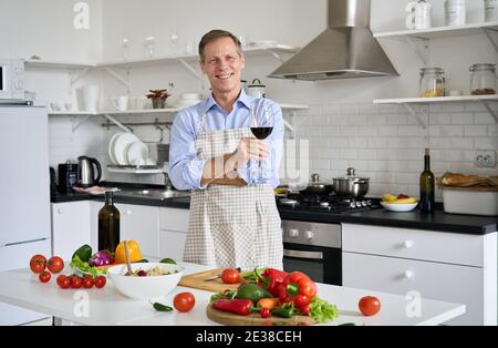 Ein glücklicher älterer Koch, der Weinglas in der Küche hält und Salat kocht. Stockfoto