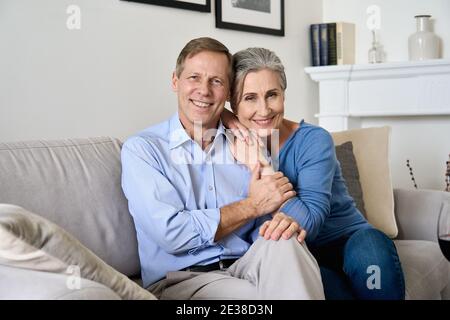 Glückliches älteres Paar umarmt, Blick auf die Kamera, sitzen auf der Couch in der Wohnung. Stockfoto