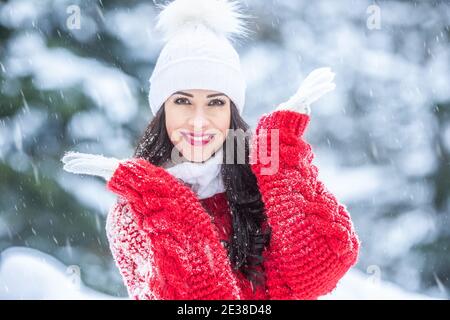 Junge Frau lächelt im verschneiten Wald und trägt Mode wie roten Pullover, weiße Handschuhe und Pom Pom Hut. Stockfoto