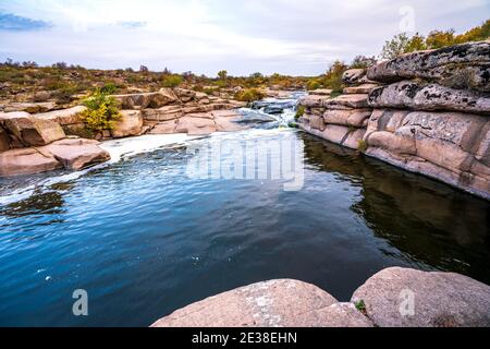 Ein kleiner, glänzender Strom fließt zwischen glattem, nassem und dunklem Wasser Steine und trockene niedrige Bäume Stockfoto