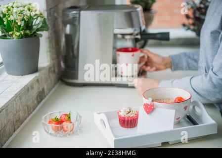 Ein Tablett mit Haferbrei Haferbrei mit Erdbeeren, Cupcake und leere Grußkarte mit Hintergrund einer Frau, die während der Zubereitung Kaffee auf der Maschine macht Stockfoto