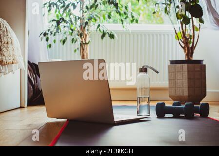 Eine Glasflasche, Laptop, Yogamatte und Kurzhanteln auf dem Boden im Schlafzimmer für das Heimtrainieren. Home Online-Workout-Konzept. Fitness online Stockfoto