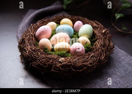 Ein Weidennest mit Moos und farbigen Ostereiern auf grauer Leinenserviette und wenigen grünen Blättern auf dunklem Grund. Dunkle Stimmung. Frohe Ostern Stockfoto