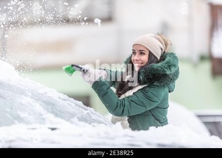Junge Frau in Winterkleidung reinigt Auto von Schnee außerhalb des Wohnblocks im Winter. Stockfoto