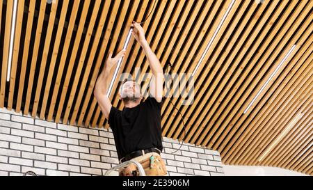 Ein Elektriker installiert elektrische Leitungen im Haus. Installation einer modernen Decke aus Holzbohlen in einem Loft-Stil Interieur. Leuchten einbauen Stockfoto