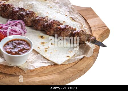 Gehackter Kebab auf einem Metallspieß mit Sauce, in Scheiben geschnittene Gurken, Tomaten und Zwiebeln auf einem Holztablett auf weißem, isoliertem Hintergrund. Stockfoto