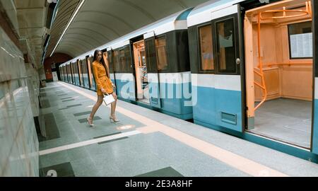 Posiert vor einer offenen Tür eines leeren U-Bahn-Zug, schöne junge Mädchen mit langen schönen Beinen in einem gelben Frühlingsmantel und einer weißen Handtasche oder Stockfoto