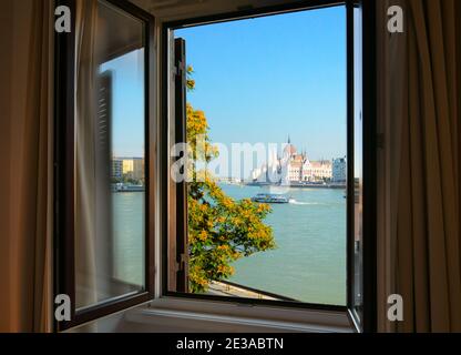 Blick auf das ungarische Parlamentsgebäude von der anderen Seite der Donau aus durch ein offenes Fenster in Budapest, Ungarn. Stockfoto