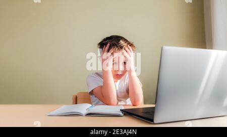 Desperate Child sitzt an einem Schreibtisch, schaut traurig auf den Computer und hält seinen Kopf mit den Händen. Problemkonzept Online-Lernen Stockfoto