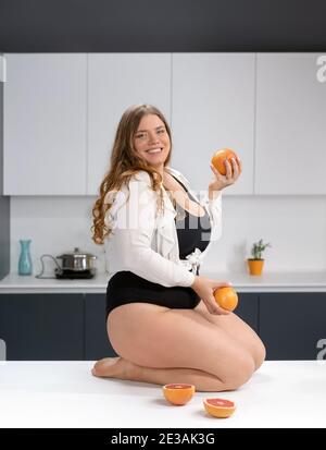 Gesundes Essen glücklich kurvigen Körper junge Frau mit langen blonden Haaren jonglieren Früchte sitzen auf modernen Küchentisch, Mischen von frischen Früchten für gesunde Stockfoto