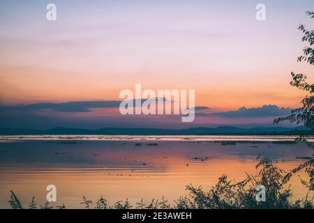 Lange Belichtung Vanille bunten Himmel und See Reflexion mit Bergen im Hintergrund. Verschwommenes wildes Gras weht im Wind im Vordergrund Stockfoto