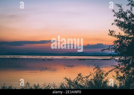 Lange Belichtung Vanille bunten Himmel und See Reflexion mit Bergen im Hintergrund. Verschwommenes wildes Gras weht im Wind im Vordergrund Stockfoto