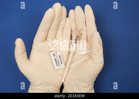 Auf blauem Hintergrund liegen zwei Fläschchen des Covid-19-Impfstoffs und eine Spritze in den Händen von medizinischen Handschuhen. Nahaufnahme Stockfoto