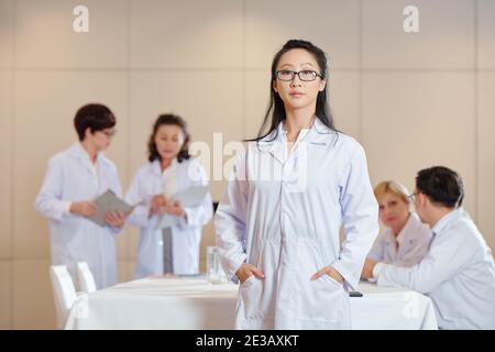 Porträt einer jungen ernsthaften chinesischen Forscherin in weißem Laborkittel, die im Konferenzraum steht und ihre Kollegen im Hintergrund sprechen Stockfoto