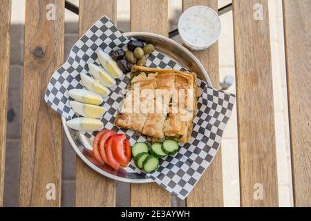 Draufsicht auf einen Teller, mit hausgemachtem traditionellen türkischen Stil Tabla Borek, gekochte Eier, Joghurt, geschnittenes Gemüse und Oliven, über einem schwarzen und weißen pa Stockfoto