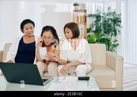 Gruppe von älteren vietnamesischen Frauen überprüfen und diskutieren Rechnungen und Online-Zahlung über Laptop Stockfoto