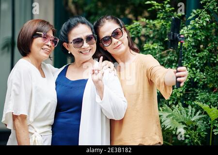 Fröhliche ältere asiatische Frauen in Sonnenbrillen posiert für Selfie auf Smartphone im Freien Stockfoto