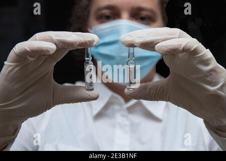 Zwei Fläschchen mit Covid-19-Impfstoff in den Händen einer Frau in einem Morgenmantel, einer Maske und Handschuhen. Nahaufnahme Stockfoto