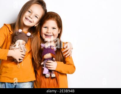 Lächelnd kleine hübsche Mädchen Schwestern in Terracotta gelb bequeme Langarm stehend umarmend halten Spielzeug Puppen, kopieren Raum Stockfoto