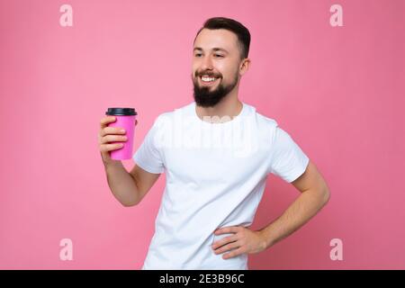 Gut aussehend positive junge Brünette bärtigen Mann trägt weißes T-Shirt isoliert Über rosa Hintergrund Wand hält Papier Kaffee Tasse trinken und Auf Stockfoto