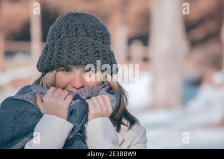 Porträt eines erröteten Mädchens, das ihr Gesicht mit ihrem Schal vor der Kälte lächelt und an einem verschneiten Tag vor der Kamera posiert. Winter Im Freien 2021. Stockfoto