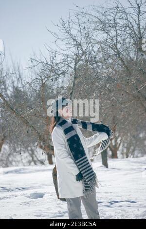 Vertikale Fotografie einer jungen Frau mit Winterhut und Handschuhen, setzt ihren schwarzen Schal an einem verschneiten Tag 2021. Winterlandschaft 2021. Stockfoto