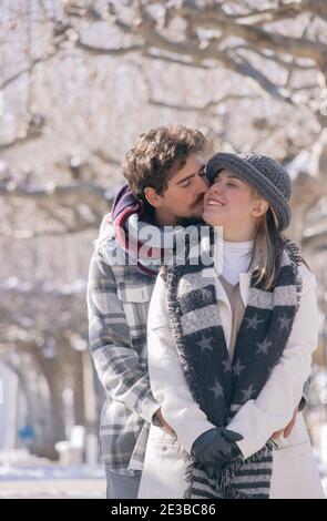 Porträt eines glücklichen Paares, gibt er ihr einen Kuss auf die Wange, während die Freundin ihn an einem verschneiten Tag fröhlich anlächelt. Winter Im Freien 2021. Stockfoto