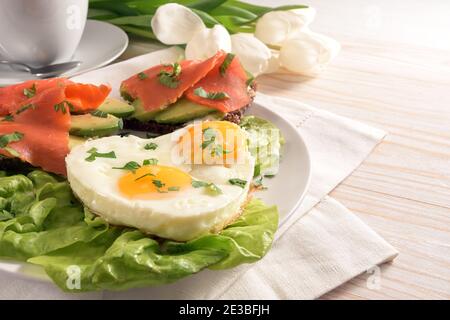 Holiday Frühstück mit Liebe, Spiegeleier in Herzform, Sandwiches mit Avocado und geräuchertem Lachs auf Salat, weiße Tulpen auf einem hell bemalten Holz t Stockfoto