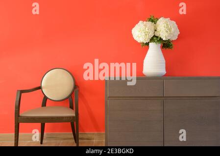 Minimalistisches Interieur mit roter Wand und künstliche weiße Blumen oder Nelken in weißer Vase & moderner Holzstuhl Stockfoto