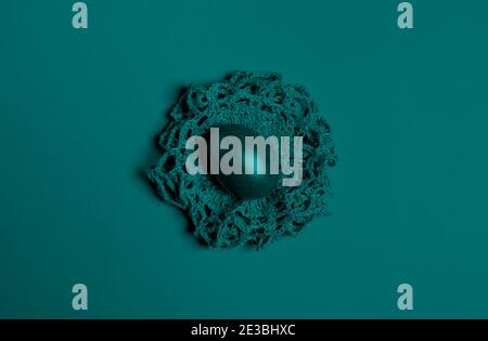 Ein Osterei liegt auf einem selbstgemachten Stricknest. Bild getönt in trendiger Farbe 2021 Tidewater Green. Kreative farbige minimalistische Osterhintergrund. Draufsicht Stockfoto