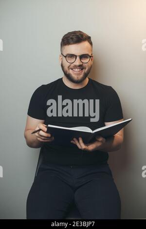 Bärtiger Mann lächelt an der Kamera an einer grauen Studiowand Ein Buch halten und eine Brille tragen Stockfoto