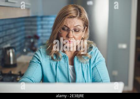 Ältere blonde Frau ist konzentriert arbeiten zu Hause bei der Laptop aus der Ferne mit Brille in der Küche Stockfoto