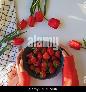 Hände der Frau halten eine Schüssel mit frischen Erdbeeren auf dem Tisch mit roten Tulpen Stockfoto